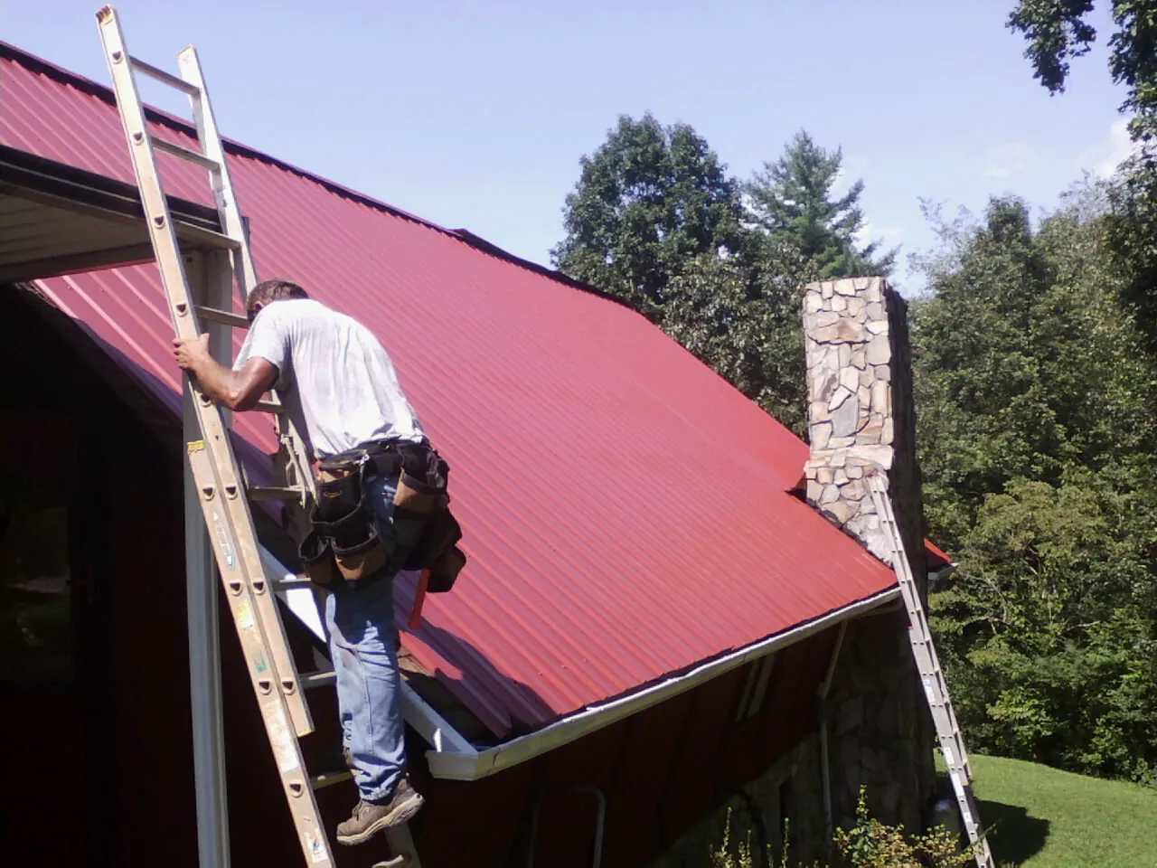 Roofer on a ladder beside a red metal roof and stone chimney — Soffit & Fascia Repair by Roofing Brothers in Bearsville