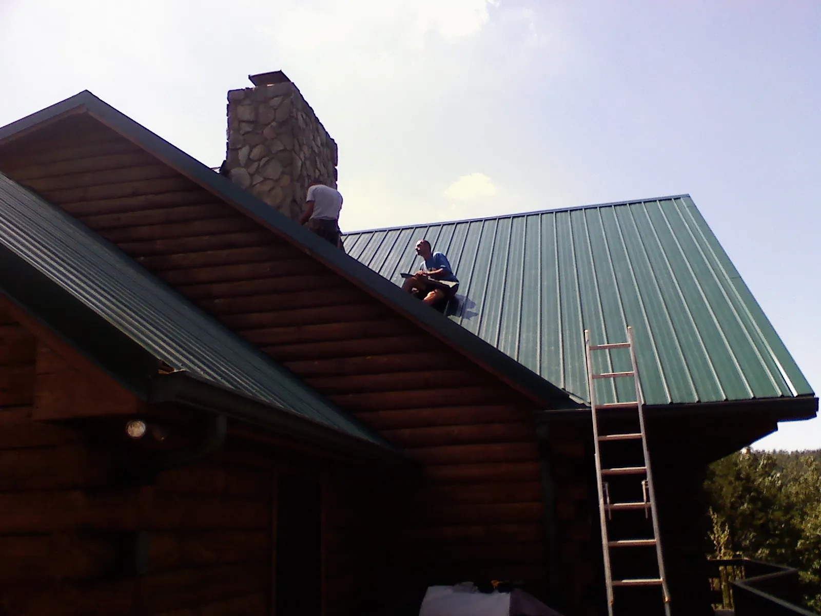 Roofers installing green metal panels beside a stone chimney on a log home in Bearsville